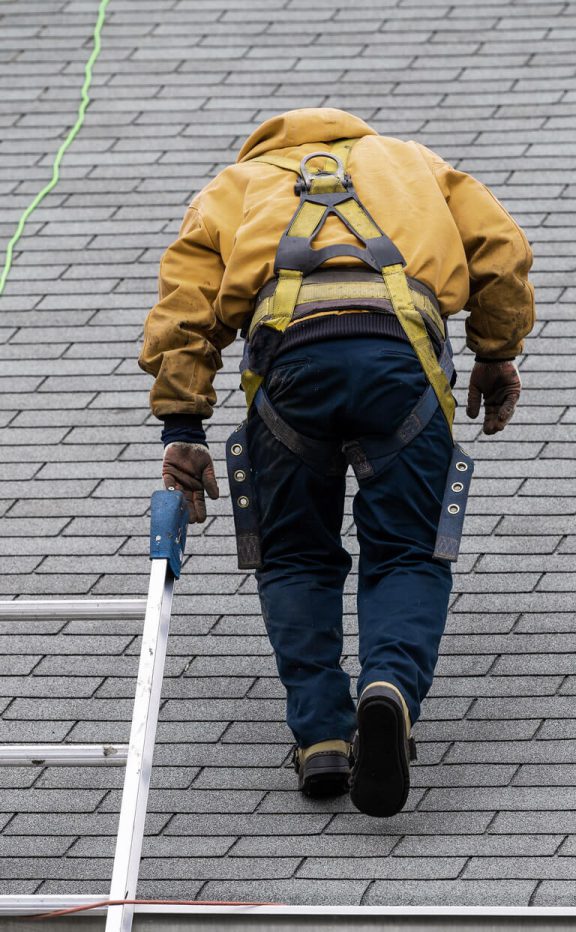 A roofer in a yellow coat climbs up on a shingled roof.